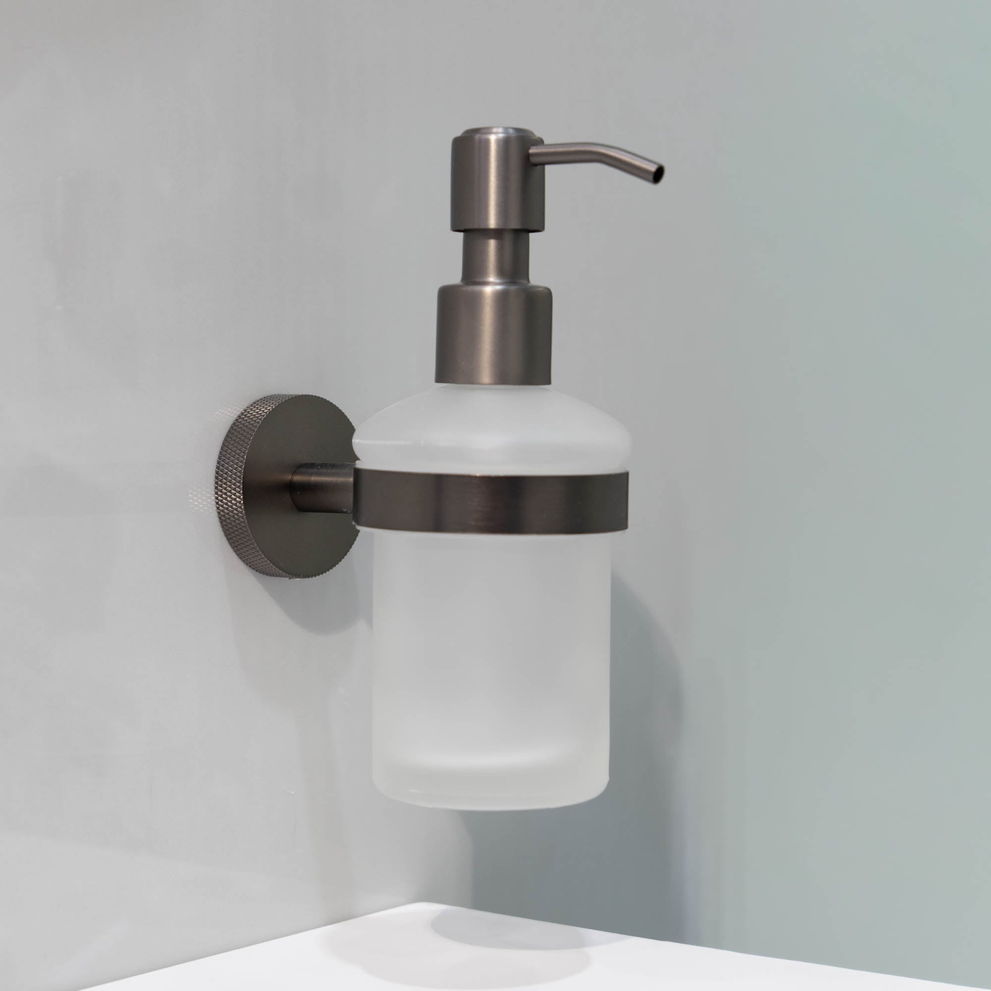 Wall-mounted soap dispenser with frosted glass container and metallic pump on a light gray background.