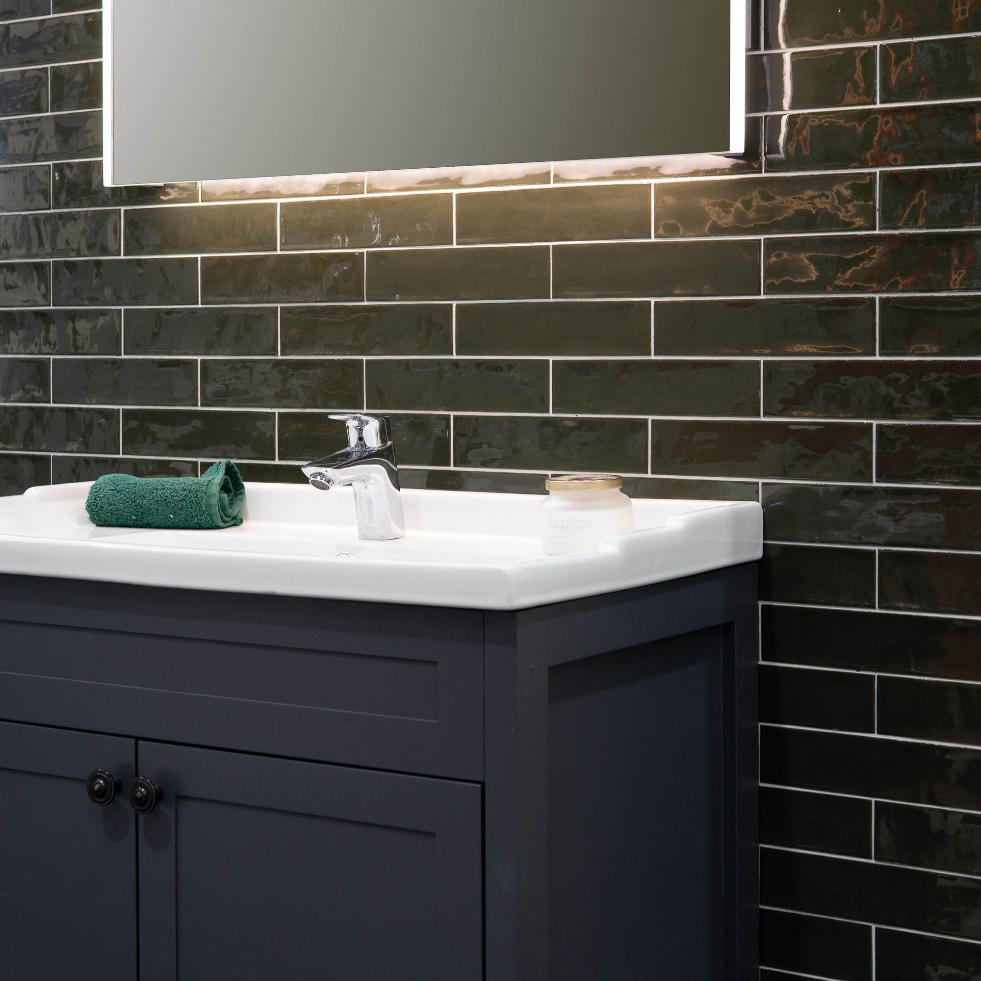 A traditional bathroom vanity with a white ceramic sink basin, chrome tap, illuminated mirror and a glossy ark green subway tile backsplash.