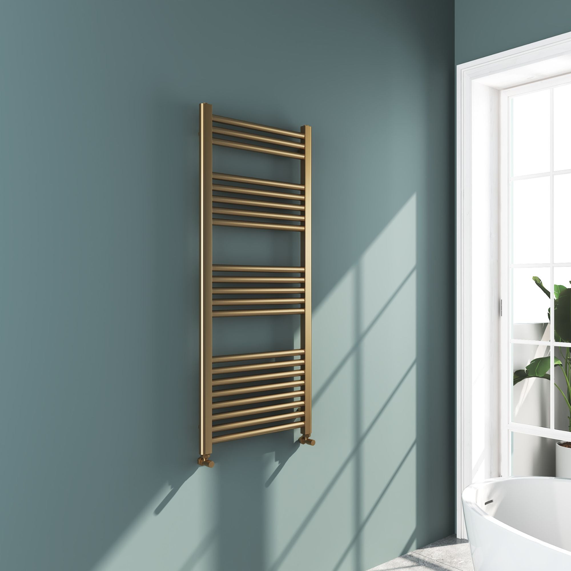 Soho brushed brass 1200x500mm towel radiator on a light blue wall with a window in the background deluxe bathrooms