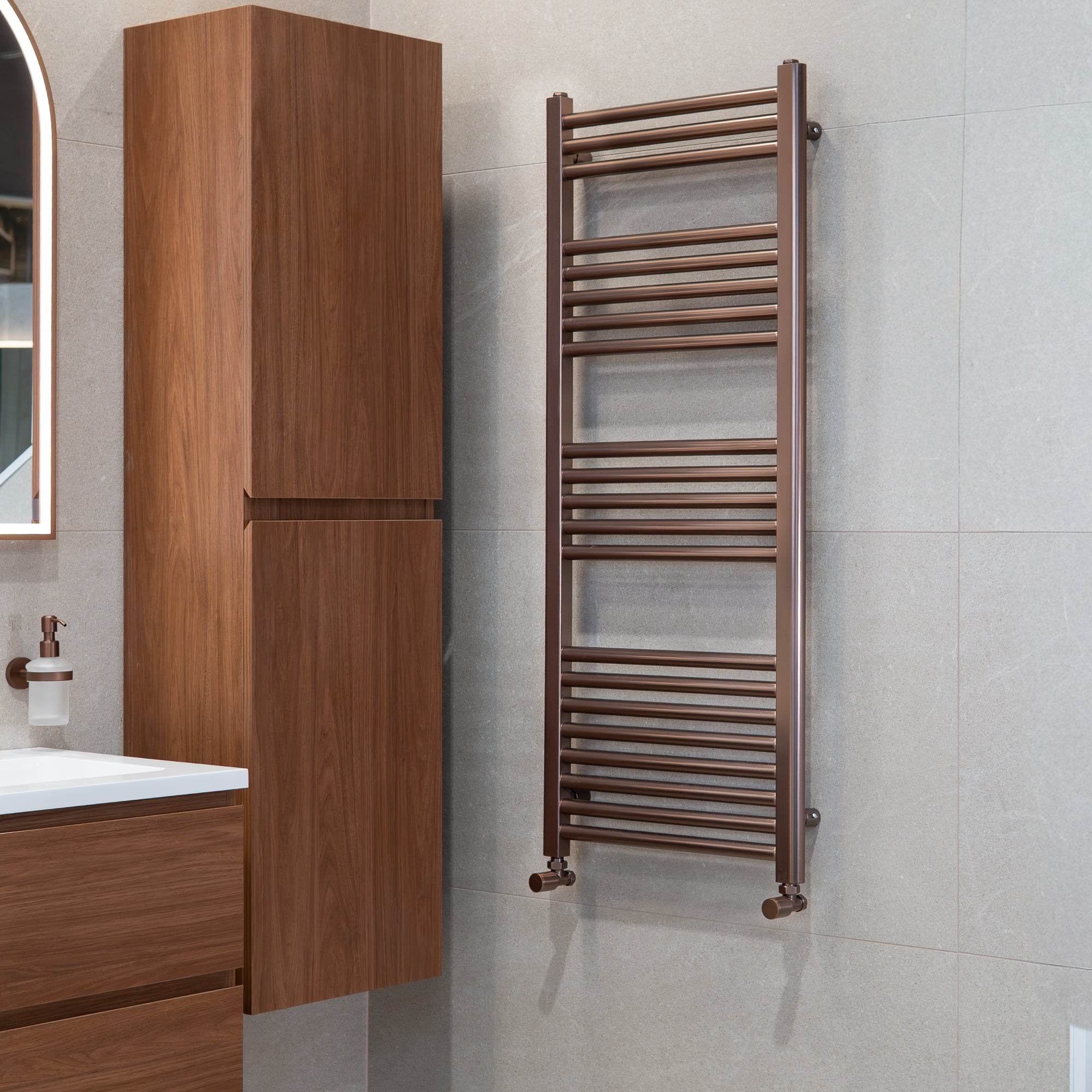Soho brushed bronze 1200x500mm towel radiator on a grey wall with a window in the background deluxe bathrooms